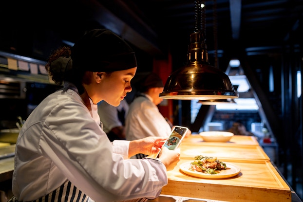 A woman in a restaurant kitchen uses a smartphone to take a picture of a meat-and-veggie dish. The kitchen is lit only by overhead lights aimed directly at the wooden counter where the dish is laid out. The woman is wearing a white chef's jacket and a black hair wrap.