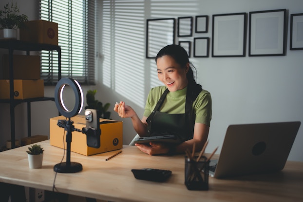 A smiling young woman sits at a table and speaks while facing a smartphone attached to a freestanding ring light. The woman holds a digital tablet and digital pen, and she wears a light green T-shirt and black apron.