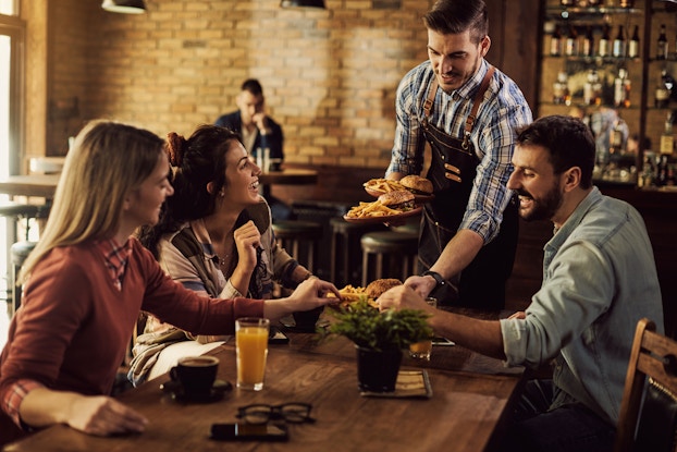 A waiter brings plates of burgers and fries to a table of three in a restaurant. The waiter is a man weraing a blue-and-white plaid shirt under a dark blue apron. He is setting one plate of food down on the table and balancing the other two plates on his arm. The three people at the table are two women sitting on the left side of the table and one man across from them on the right. One of the women and the man are reaching for fries on the plate being lowered in front of them. The other woman looks up at the waiter with a smile.