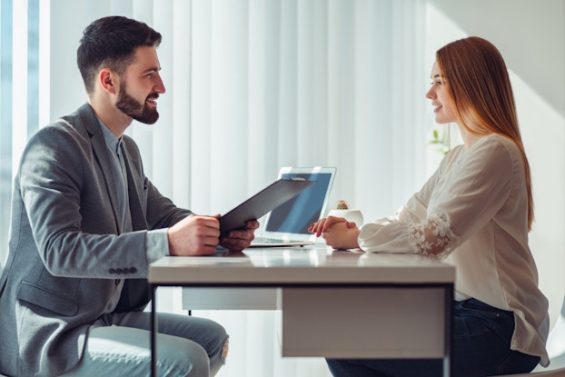 A profile shot of a man and woman sitting on opposite sides of a metal desk and smiling at each other. The man is seated on the left; he has dark hair and a dark beard. He wears a gray blazer and holds a clipboard. The woman is seated on the right; she has long strawberry blonde hair and wears a white blouse with a partially transparent panel at the elbows. She sits with her hands folded.