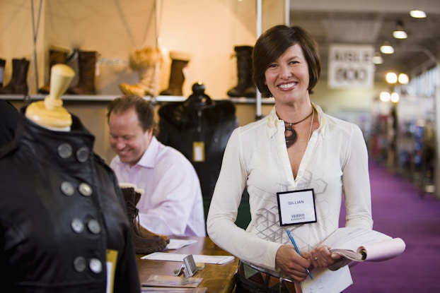 A woman stands smiling in front of a clothing company's booth at a trade show. The woman has bobbed brown hair and wears a white blouse with a hexagonal pattern across the front and a name tag with the name "Gillian" and a barcode. The clothing company's booth displays leather jackets on mannequins and leather and fur-lined boots on shelves. A man sits at a desk in the booth.