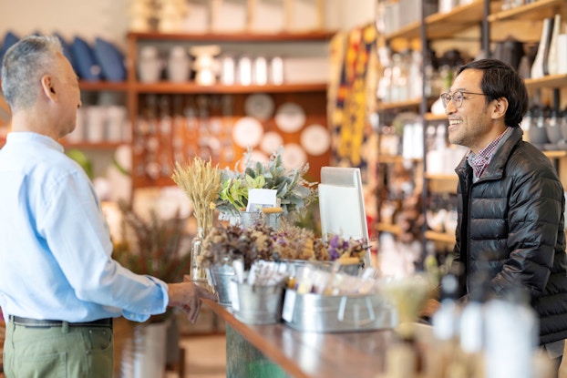 A retail shop owner talks to a customer in his shop. Both men are smiling while they are engaged in a conversation.