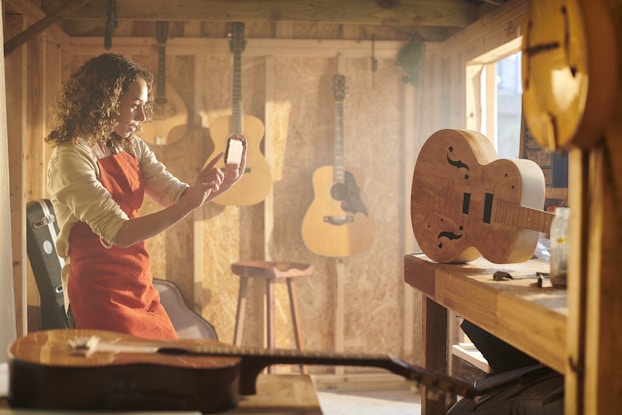 A woman in a workroom with unfinished walls uses her smartphone to take a picture of a fully carved but unstrung wooden guitar on a worktable. Finished guitars hang on the walls of the room.