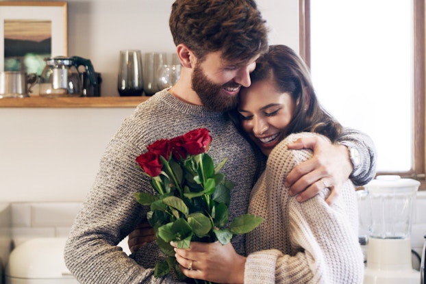 A happy couple hugging in their living room while they hold a bouquet of roses.