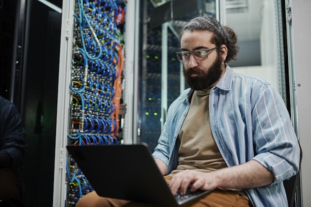 A man sits next to a server bank and types on a laptop. The man has dark hair pulled back into a man bun and a full beard. He's wearing glasses and a blue-and-white striped button-up shirt over a brown T-shirt.
