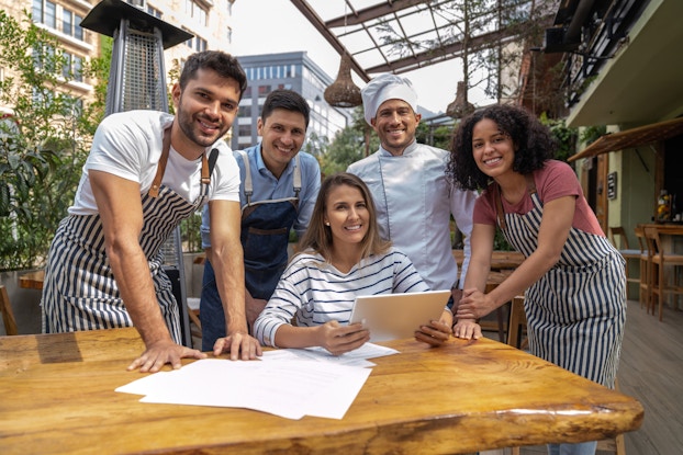 A group of restaurant workers stand around a woman seated at a table outside of a restaurant. The four restaurant employees are three men and a woman. One of the men is wearing a chef's jacket and hat; the rest of the restaurant employees are wearing aprons. The woman at the table is wearing a black-and-white-striped shirt and holding a digital tablet. Some papers are spread on the table in front of her. All five people are facing the viewer and smiling. In the background is the space outside of the restaurant, with the business's building off to one side, some trees and other buildings off to the other side, and a glass-paneled awning extending over the sidewalk above the group of people.