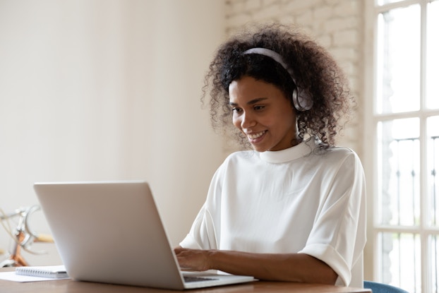 A smiling young woman sits at an open laptop with her hands on the keyboard. She wears a white shirt and silver headphones.
