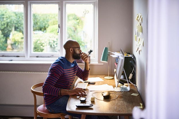 A wide shot of a bearded man sitting at a desk, looking at a computer monitor. The man is wearing glasses and a blue shirt with orange horizontal stripes. He has one hand to his mouth in a thoughtful expression and the other hand on a computer mouse. The desk also holds a lamp, a cordless keyboard, a cordless phone, a smartphone, and a stacked set of trays holding various papers.