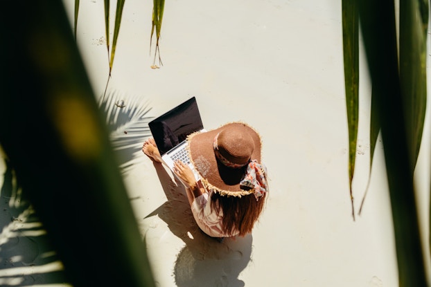 Top view of a woman working on her laptop while sitting in the sun on white sand.