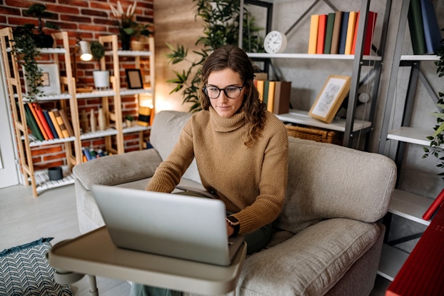 A woman wearing glasses and a tan turtleneck sweater sits in a plush beige armchair and looks at an open laptop. The laptop sits on a small plastic table in front of the chair. In the background are several shelves filled with books, framed pictures, and a few potted plants.