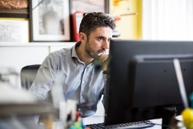 A man is seated at a desk in front of desktop computer. He has a serious look on his face. He is looking at data on his computer and thinking.