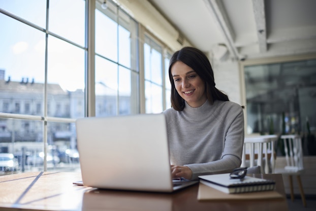 A young smiling woman with long, dark hair sits at a table and types on a laptop. She sits in a large room next to a wall of windows that look out over several buildings.