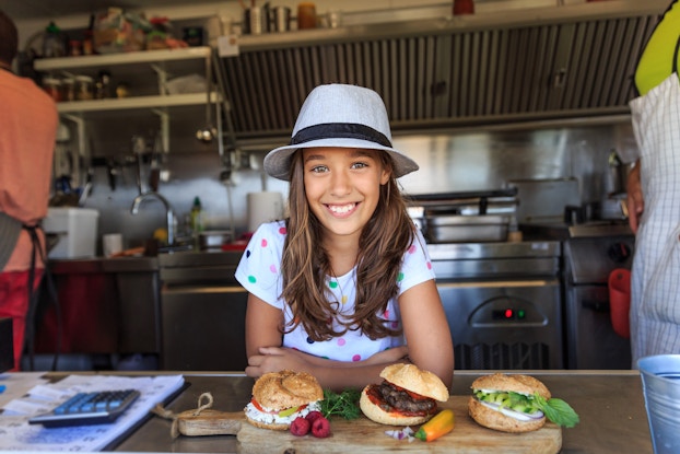 Young girl standing in a kitchen in front of three sandwiches.