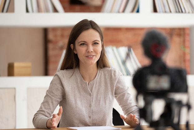 A young woman sits at a desk and speaks to a camera, which sits out of focus in the foreground.